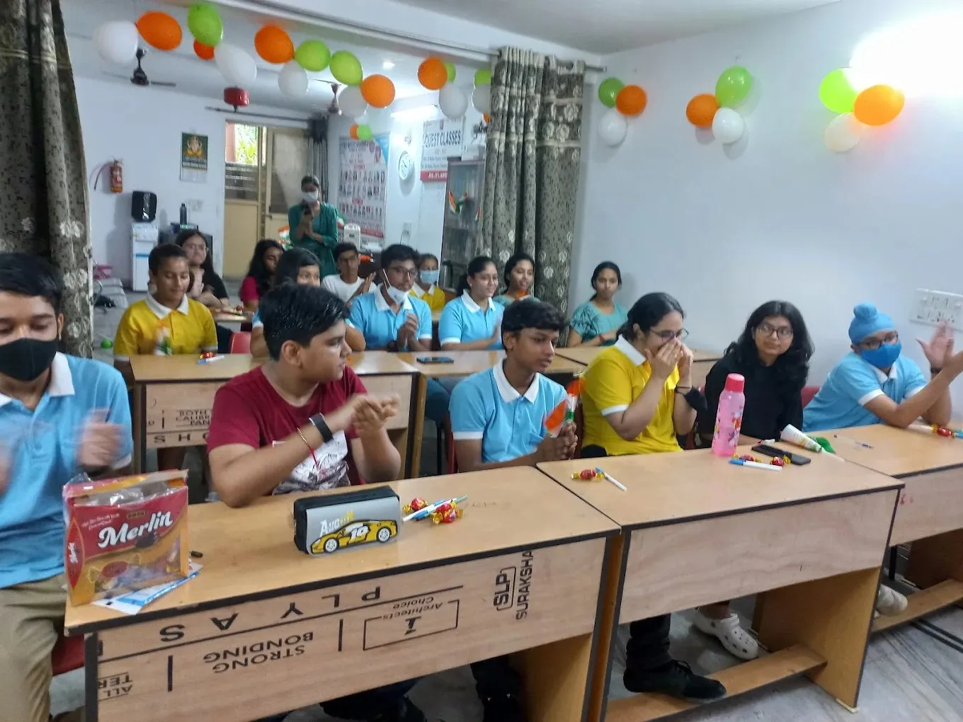 Students clapping at their desks in a room decorated with tricolour balloons.